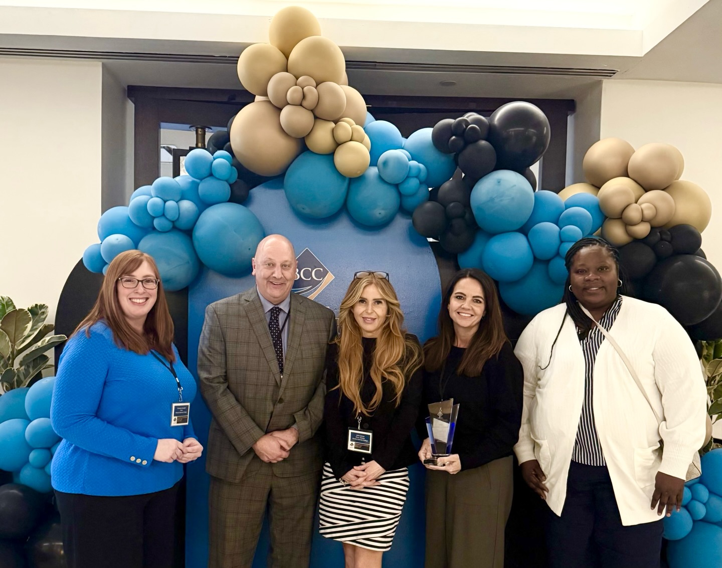 Five staff members pose with an award in front of a decorative balloon display at a formal indoor event.