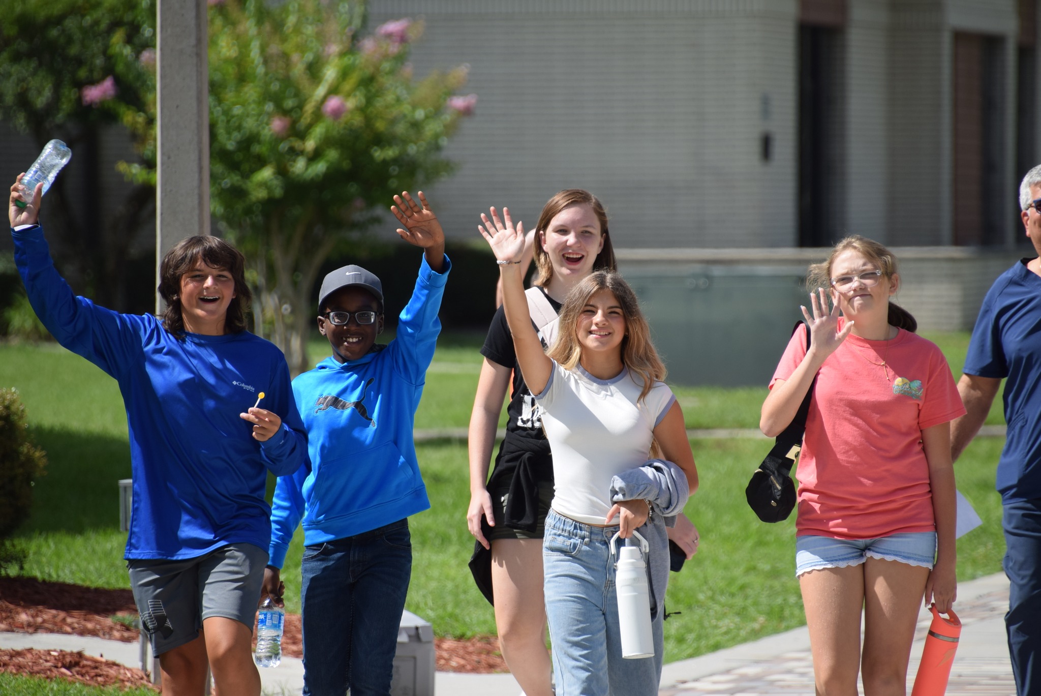 Group of campers walking outdoors on campus, smiling and waving at the camera on a sunny day.