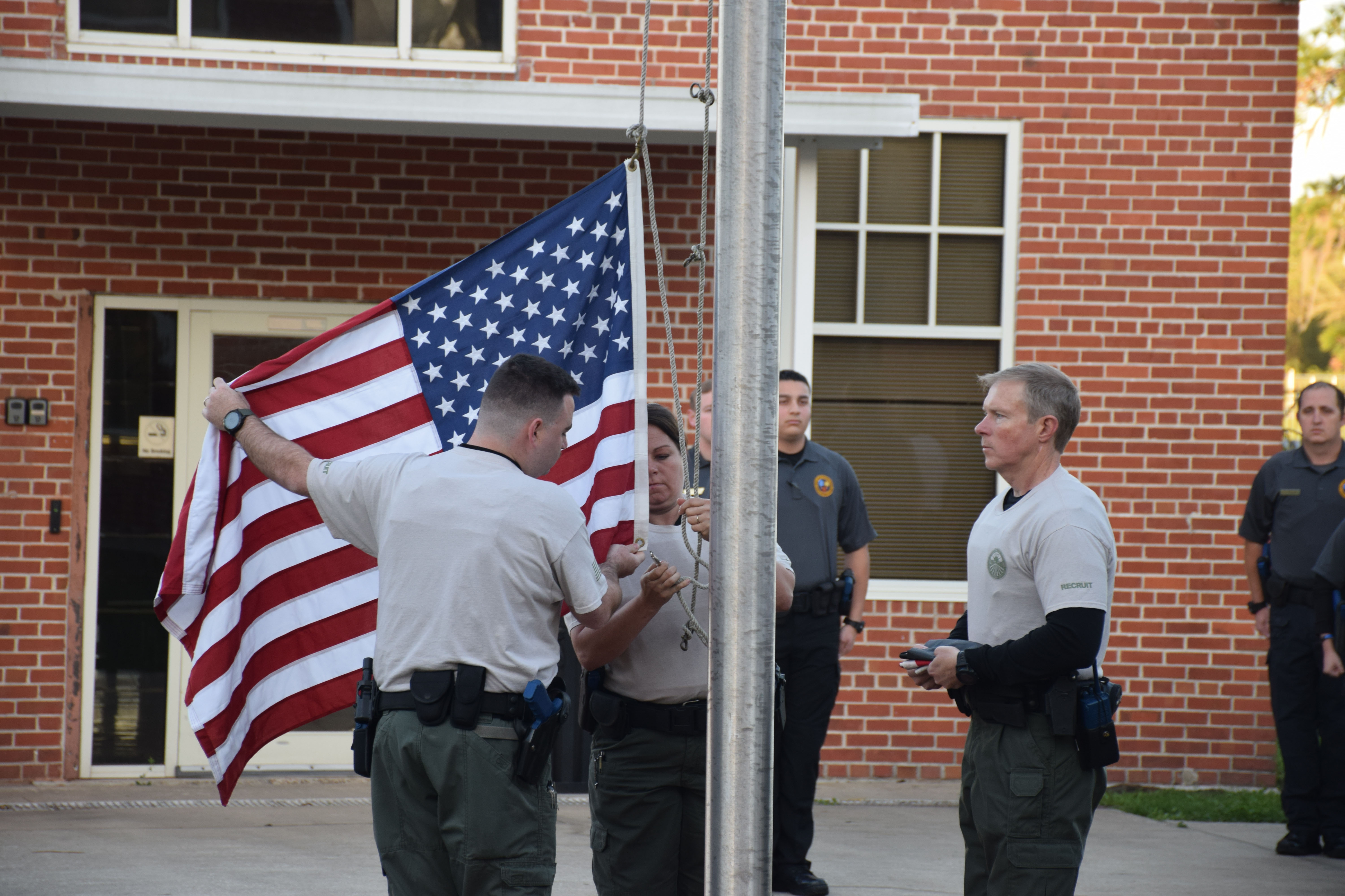 Law enforcement recruits raising the American flag on a flagpole outside a brick building, with uniformed officers standing at attention nearby.
